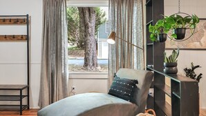 A bedroom with a bed, a chair, a desk, a hanging plant, and a window with curtains.