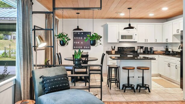 A modern kitchen with white cabinets, a black countertop, and a central island with stools. There is a dining table with chairs and a hanging plant.