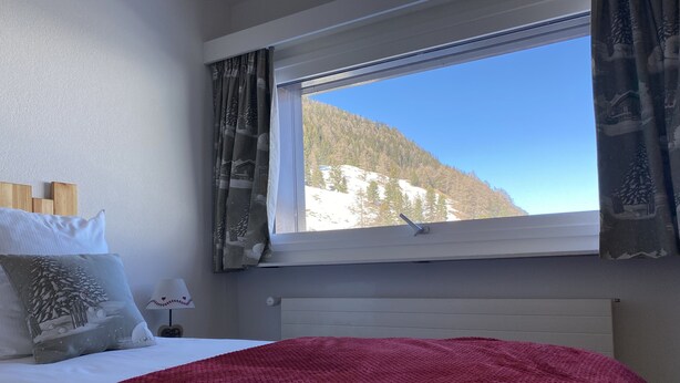 A bedroom with a bed, a window with curtains, a radiator, and a view of a snowy mountain. - La Marmotte (Siviez)