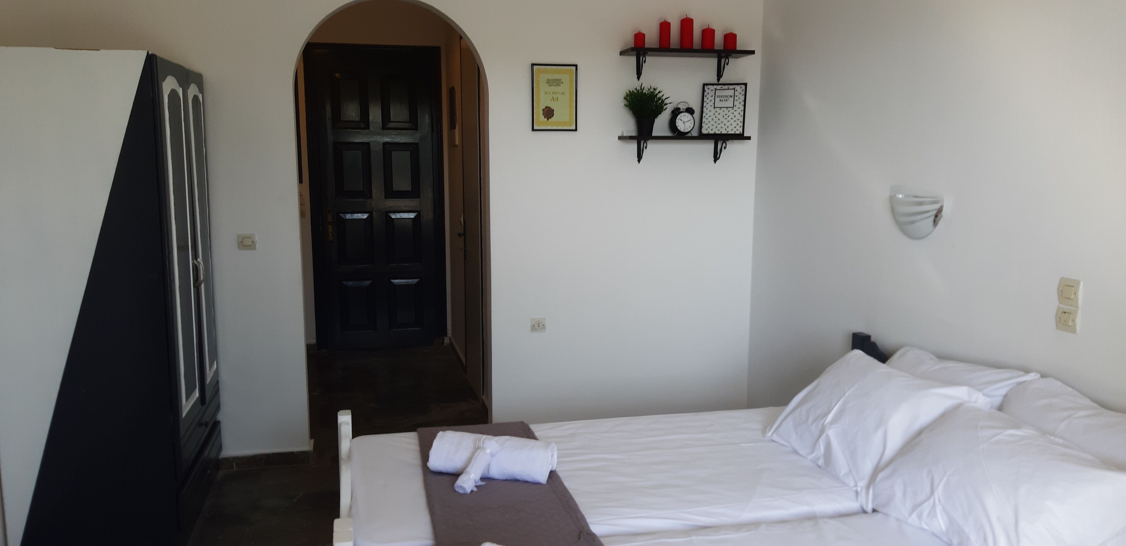 Une chambre d’hôtel avec un lit, une porte, une étagère avec des bougies et une horloge murale.