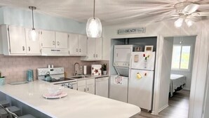 Une cuisine moderne avec des meubles blancs, un îlot central doté de tabourets de bar, un réfrigérateur orné d’aimants et un panneau indiquant « PLAGE ».