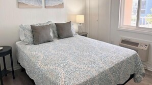 Une chambre à coucher avec une couvre-lit à motifs, deux oreillers, une table de chevet avec une lampe, une fenêtre donnant sur un paysage agréable et deux tableaux accrochés au mur.