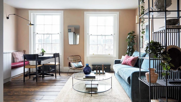 Un salon comprenant un canapé, une table basse, un fauteuil et un radiateur. Il y a deux fenêtres et un miroir au mur.