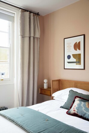 A bedroom with a bed, a window with curtains, a bedside table with a lamp, and a wall with a framed abstract artwork.