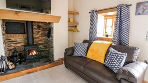 A cozy living room with a stone fireplace, a leather sofa with patterned cushions, and a large window with curtains. - The Old Cobblers Shop (Llandrindod Wells)