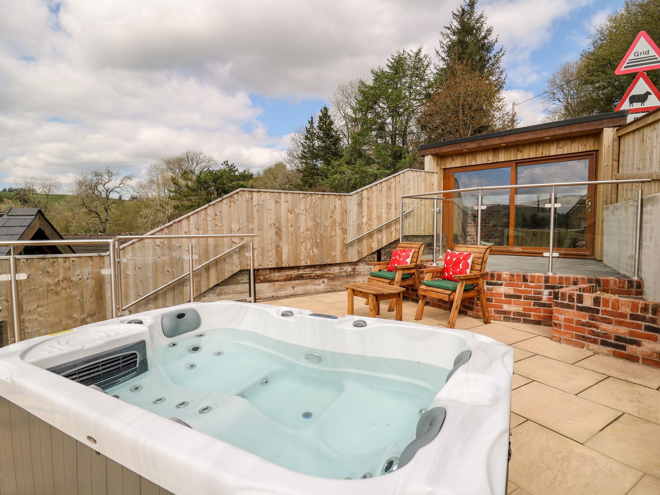 A hot tub with a wooden deck, two wooden chairs with cushions, and a brick wall.
