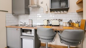A compact kitchen with a microwave, toaster, and a small refrigerator. There are two grey bar stools and a shelf with dishes above the counter. - The Old Cobblers Shop (Llandrindod Wells)