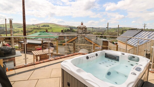 A hot tub on a balcony with a view of a green landscape and a stone building in the background. - The Old Cobblers Shop (Llandrindod Wells)