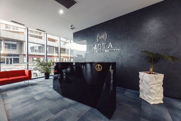 A reception area with a black reception desk, a red sofa, a potted plant, and a large window offering a view of the street. - Meta Business Hotel (Tbilisi)