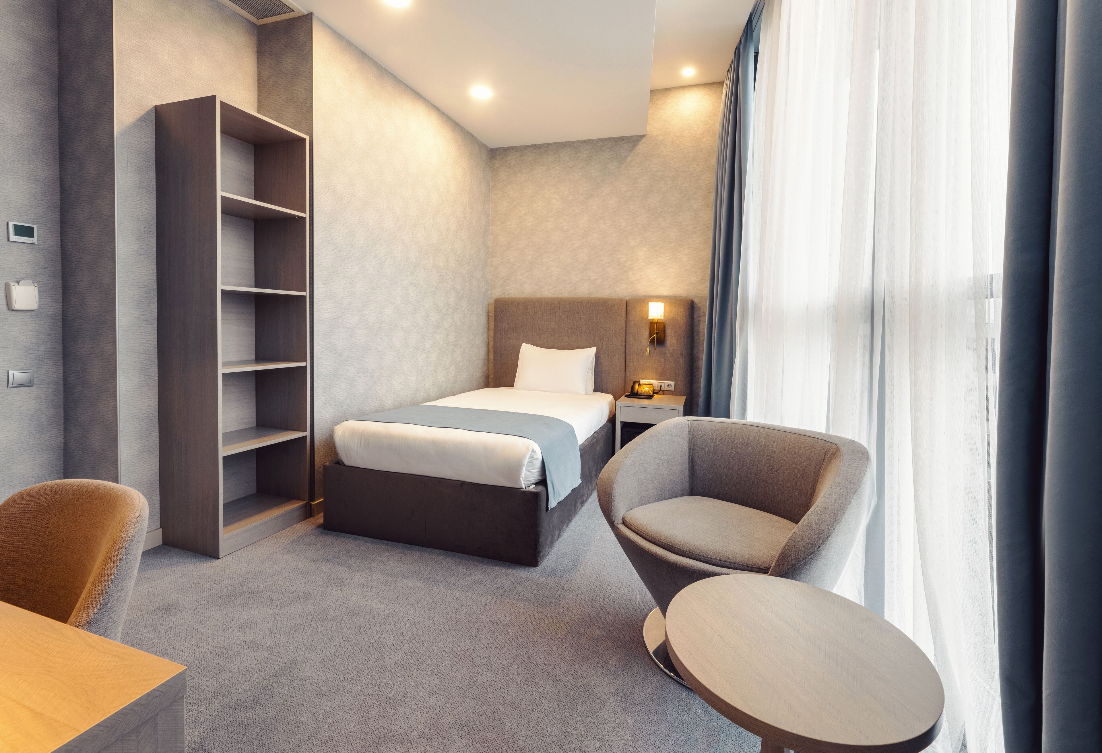 A modern hotel room with a bed, a seating area with a chair and a small table, a bookshelf, and a nightstand with a lamp.