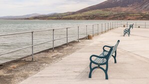 A waterfront promenade with benches and a railing overlooking a body of water and distant hills.