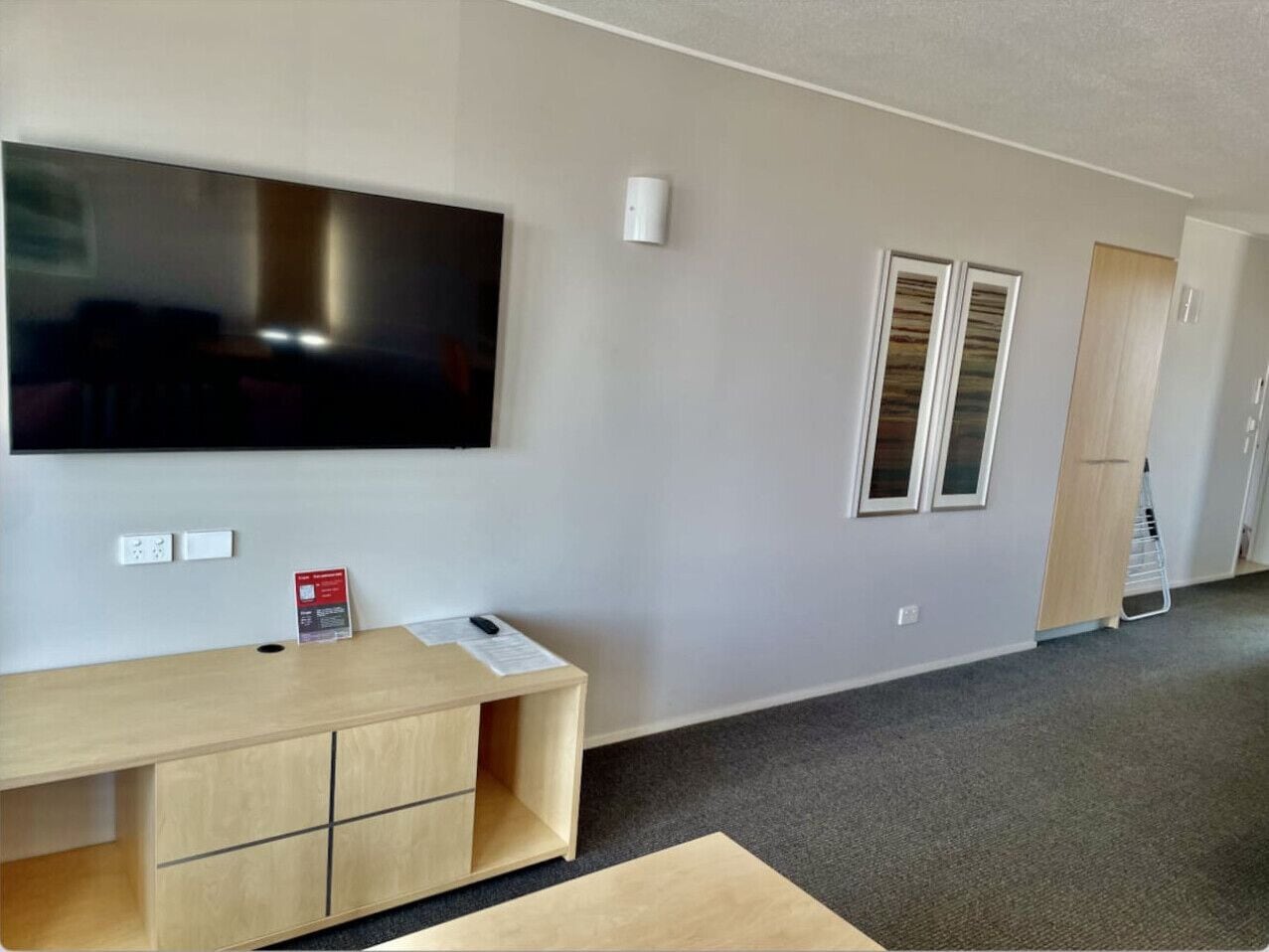 A modern room with a flat-screen TV mounted on the wall, a wooden desk with drawers, a small table with a red sign, and a window with blinds.