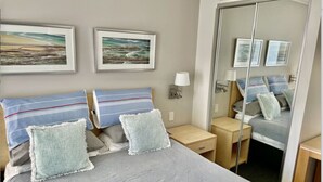 Une chambre à coucher bien rangée, avec un grand lit, deux tables de chevet, une commode, un miroir et des œuvres d’art encadrées accrochées au mur.