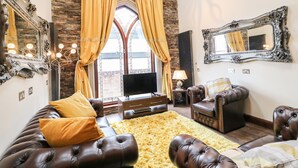 A spacious living room with a stone wall, leather sofas, a large window with yellow curtains, and a chandelier. - The Upper Room (Ballymena)