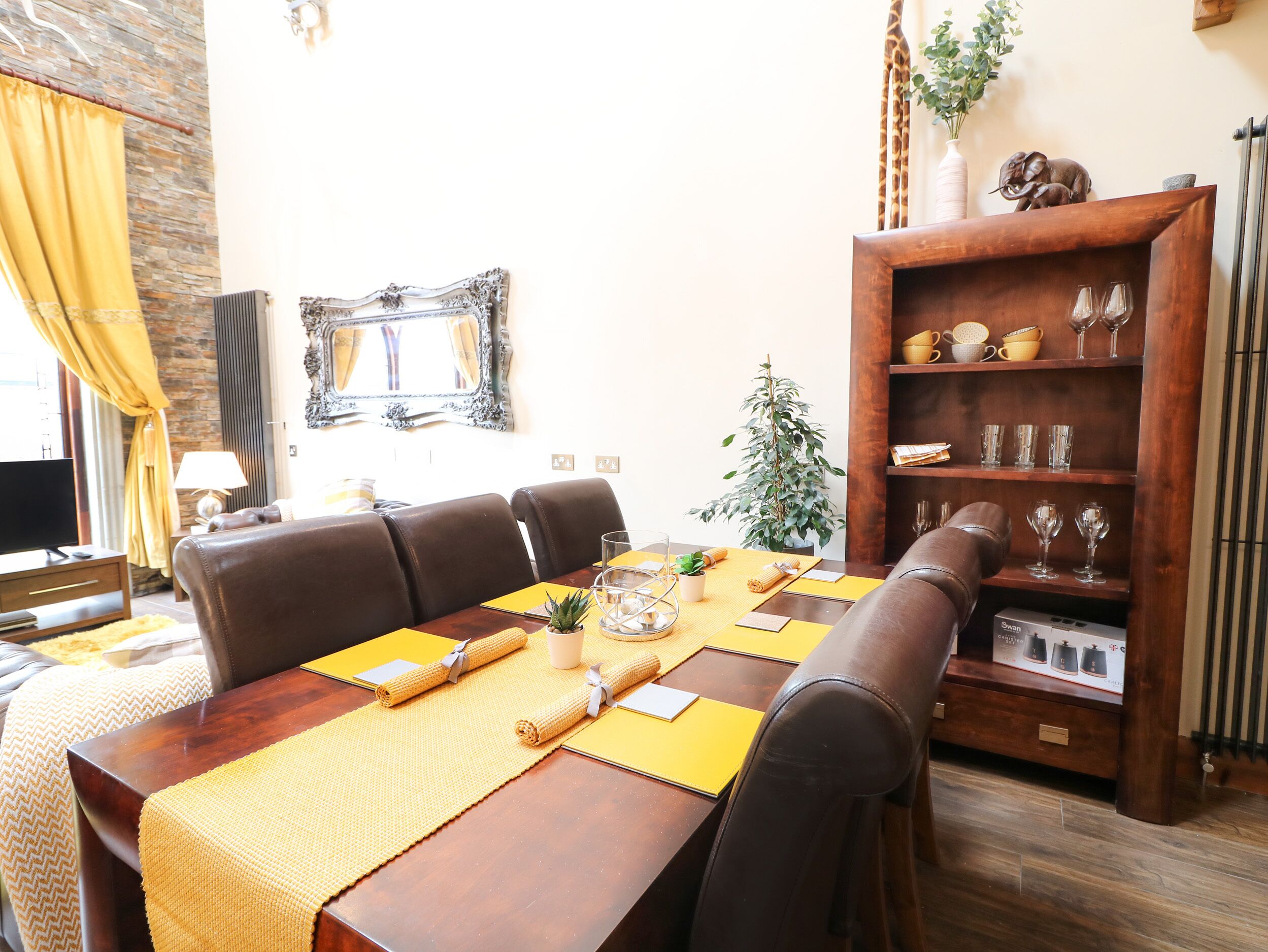 A dining area with a wooden table set for four, surrounded by leather chairs. A potted plant and a decorative shelf with glassware are visible.