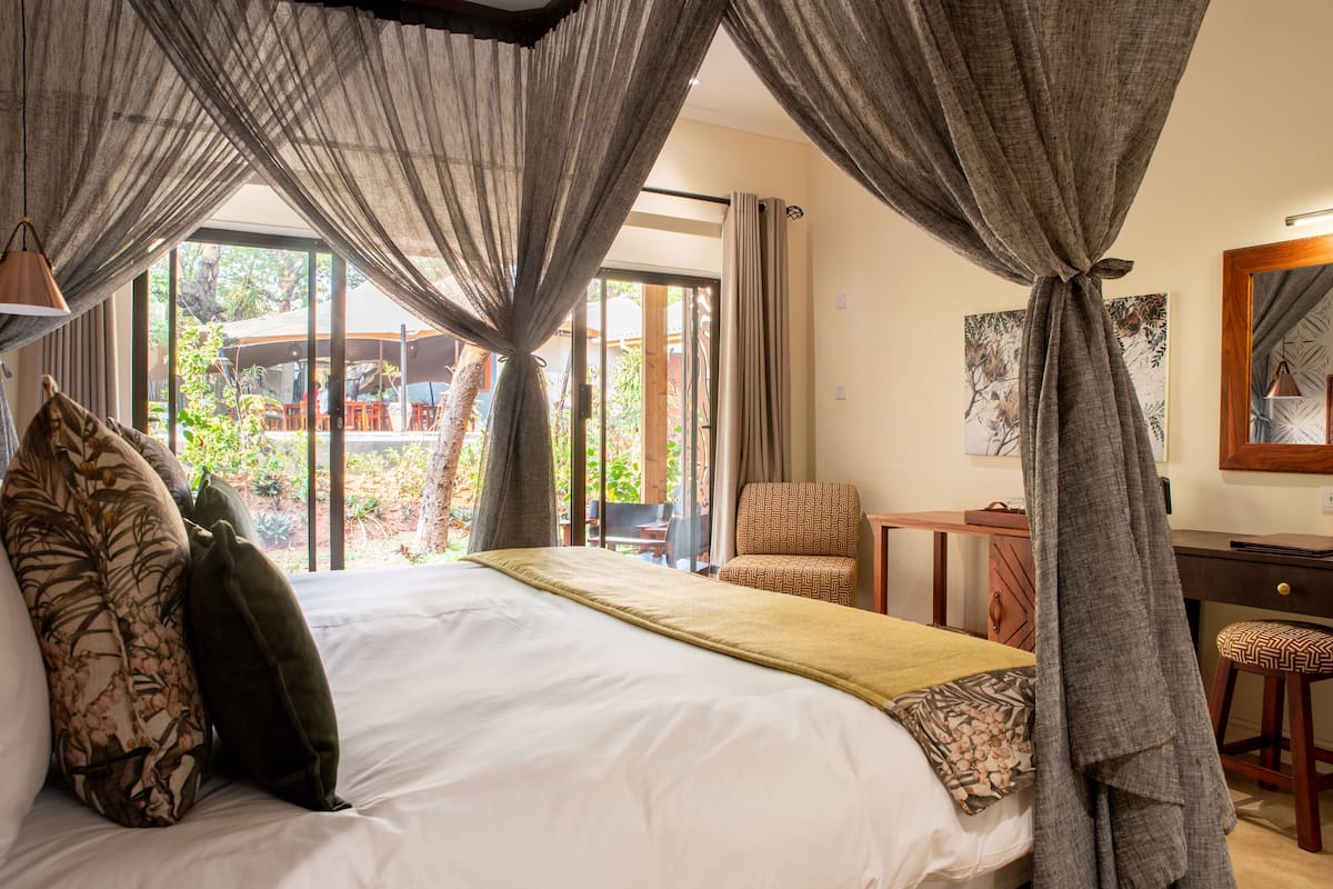 Ein Schlafzimmer mit einem großen Bett, Vorhängen, einem Sessel, einem Schreibtisch und Blick ins Freie.