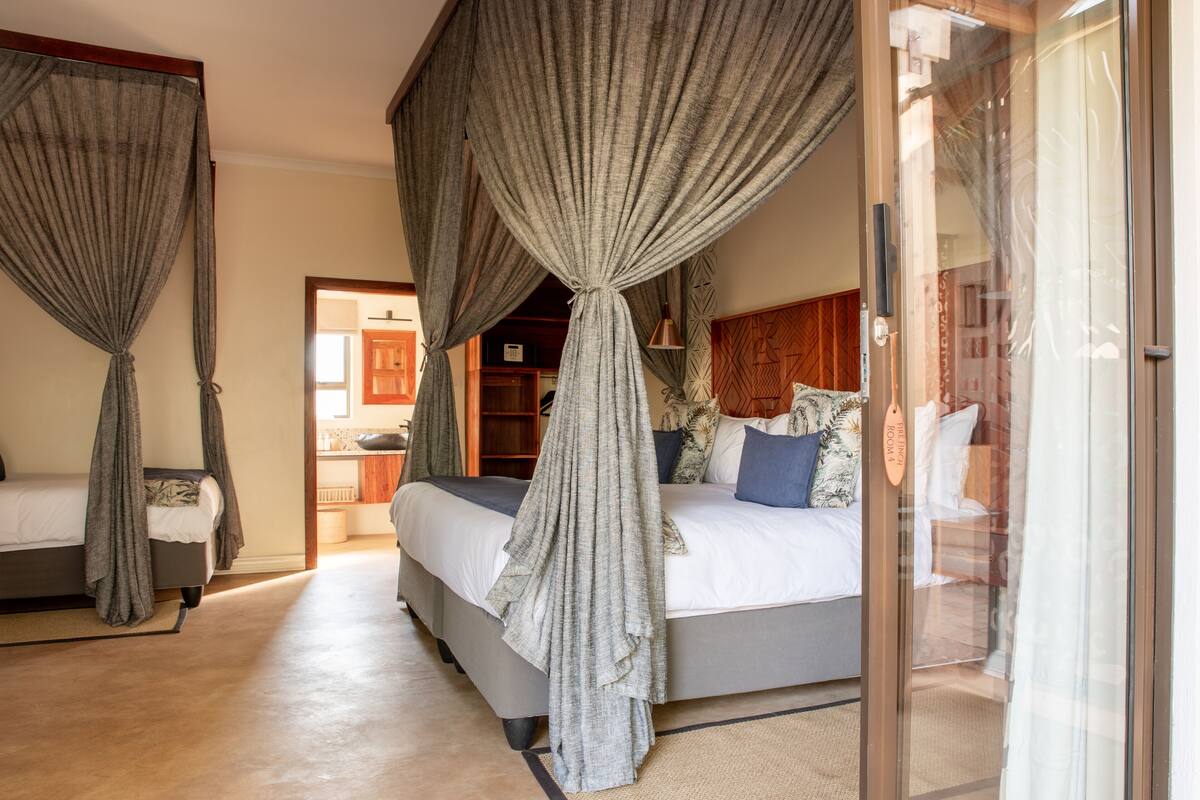 Ein Schlafzimmer mit einem großen Bett, einem kleineren Bett und einem Himmelbett. Es gibt einen Holzkopfteil und einen Nachttisch mit einer Lampe. Der Raum hat einen Holzboden und Blick ins Freie.