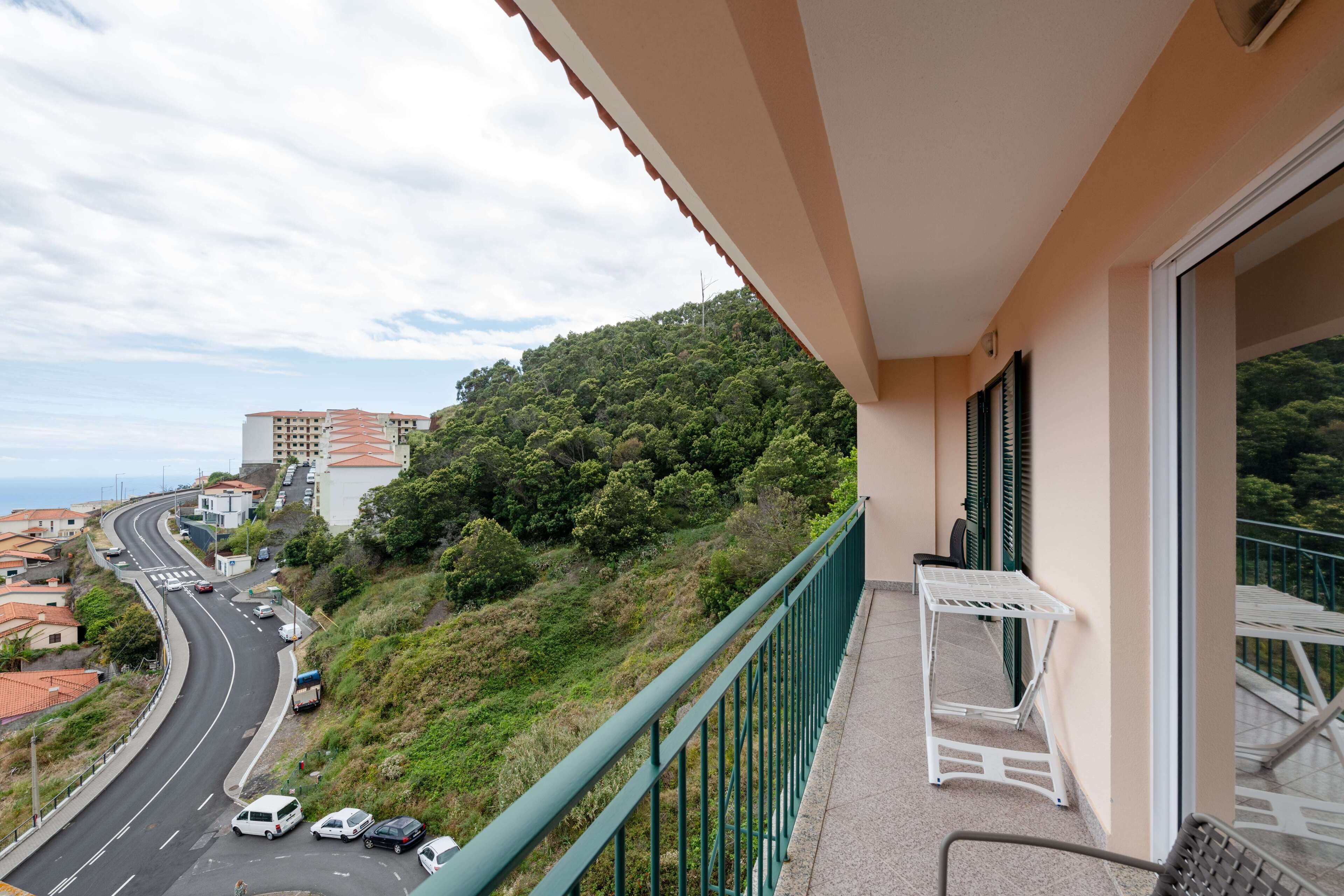 A balcony with a view of a winding road and a hill with dense greenery.