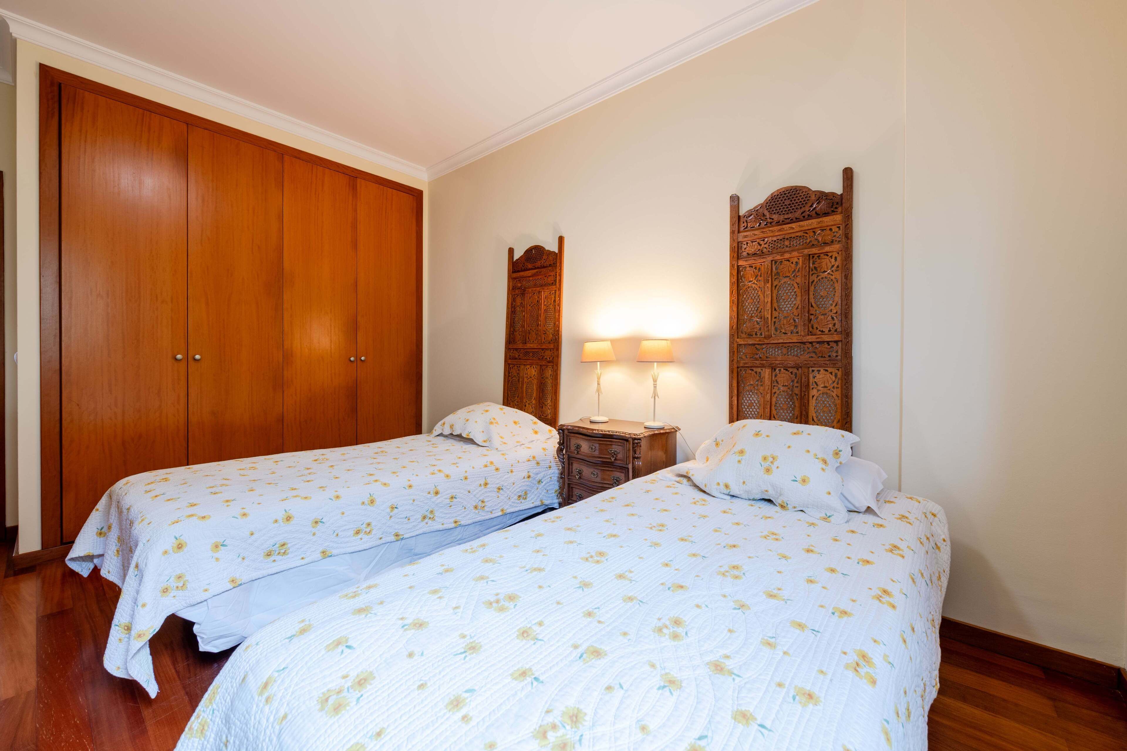 A bedroom with two single beds, wooden wardrobes, a nightstand with lamps, and a decorative headboard.