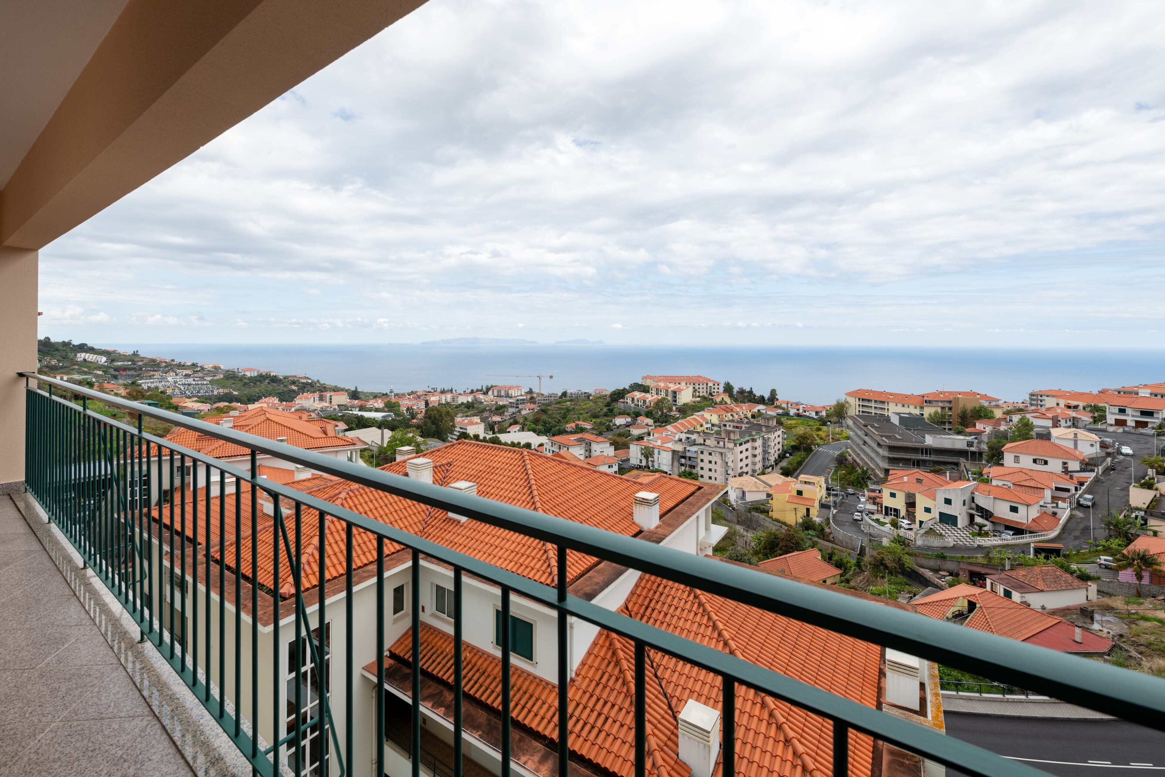 A balcony with a view of a coastal town, featuring terracotta-roofed buildings and a clear sky.