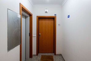 A hallway with a wooden door marked 'M', a metal door, and a blue sign indicating a men's restroom. - Caniço Quintas III (Santa Cruz)