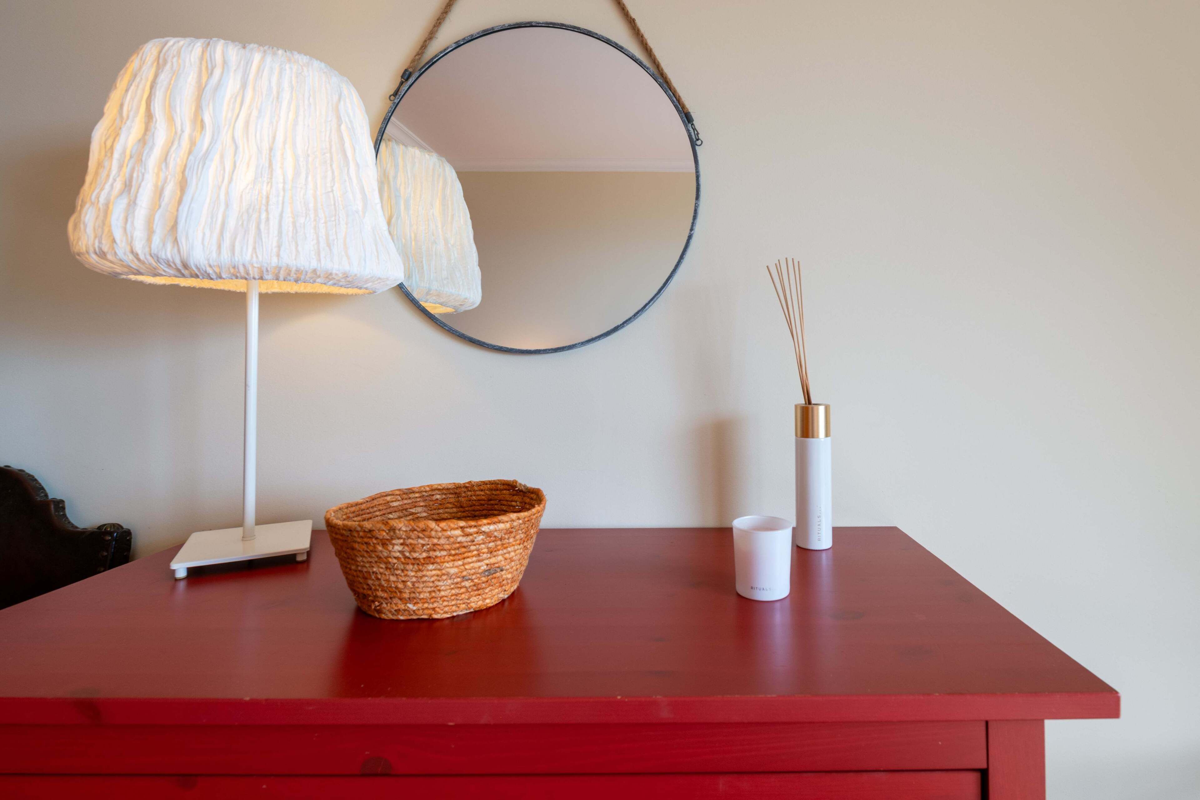 A red table with a white lamp, a woven basket, a white candle, and a mirror.