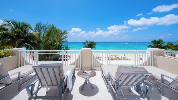 Ein Balkon mit Liegestühlen und einem kleinen Tisch, mit Blick auf einen Strand mit klarem, blauem Wasser und Palmen.