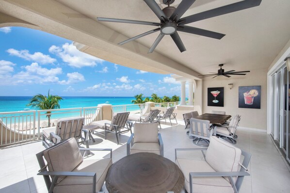 A balcony with a view of the ocean, furnished with chairs and a table, and ceiling fans. - Beachcomber 12 by Grand Cayman Villas (7 Mile Beach)