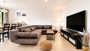 A modern living room with a sectional sofa, a flat-screen TV, a dining area with chairs and a table, and a large window. - ANDIAMO GARDENS APARTMENT (Vilamoura)