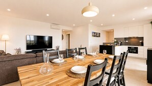 A modern living room with a wooden dining table set for four, a flat-screen TV, and a kitchen area with white cabinets and a black countertop. - ANDIAMO GARDENS APARTMENT (Vilamoura)