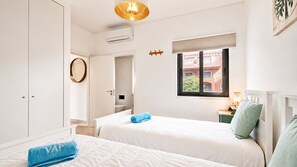 A hotel room with two beds, a window with a view of buildings, a round mirror, and a small table with a lamp.