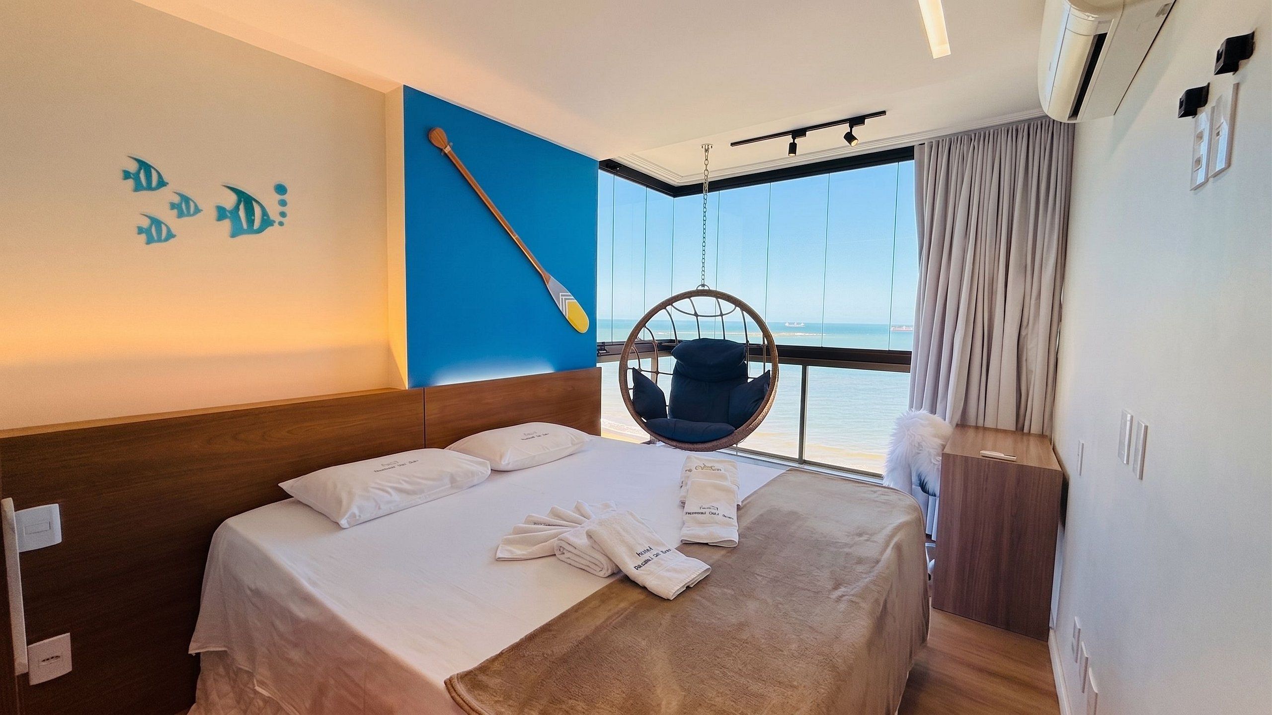 A hotel room with a bed, a hanging chair, a view of the sea, and a wall with fish decorations.