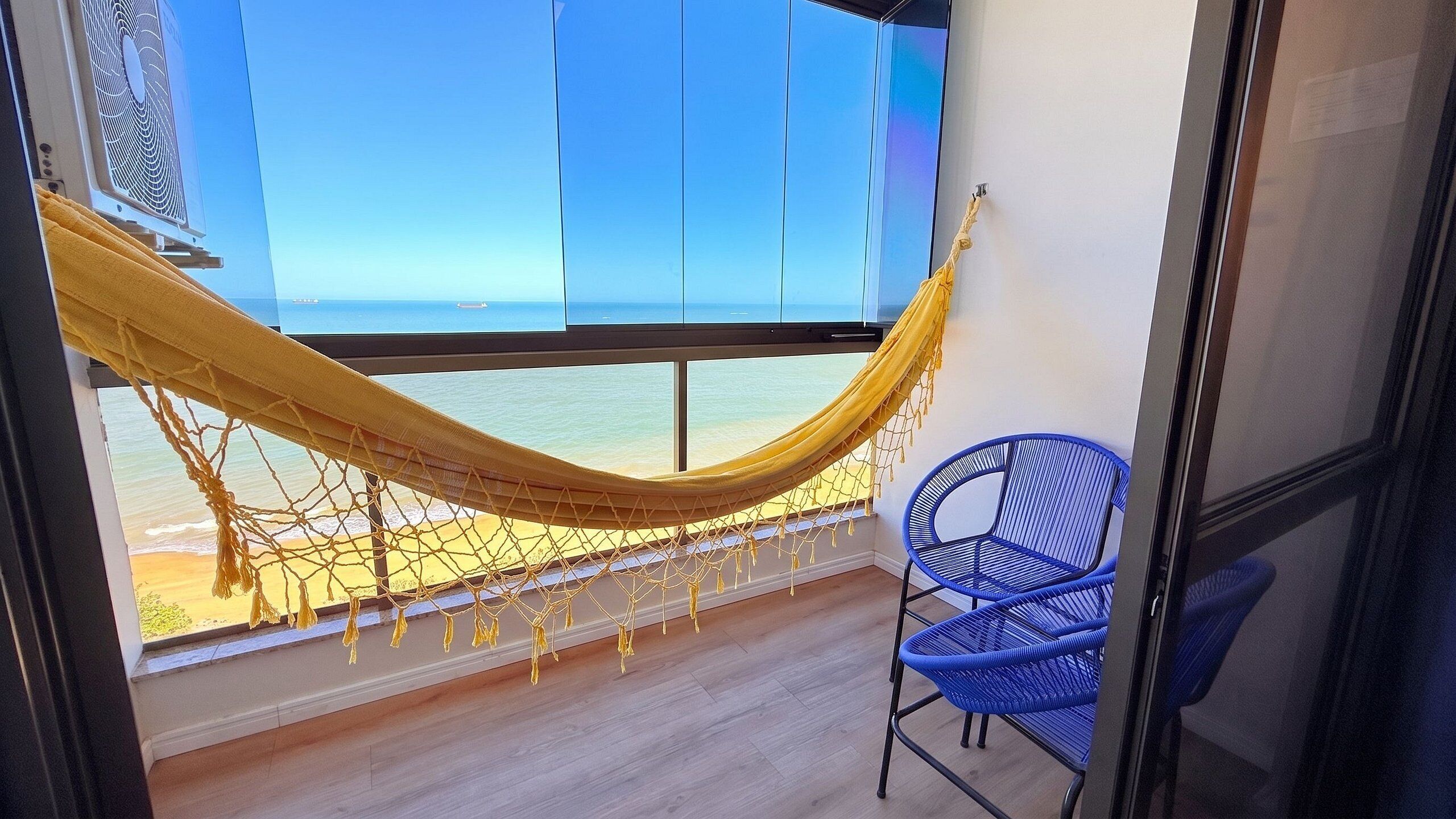 A room with a hammock, a blue chair, and a large window overlooking the sea.