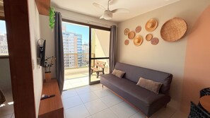 A modern living room with a grey sofa, a ceiling fan, a balcony with a view of buildings, and decorative woven items on the wall.