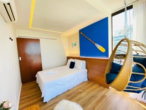 A hotel room with a bed, a hanging chair, a wall-mounted paddleboard, and a window with a city view. - Praia Dourada (Vila Velha)