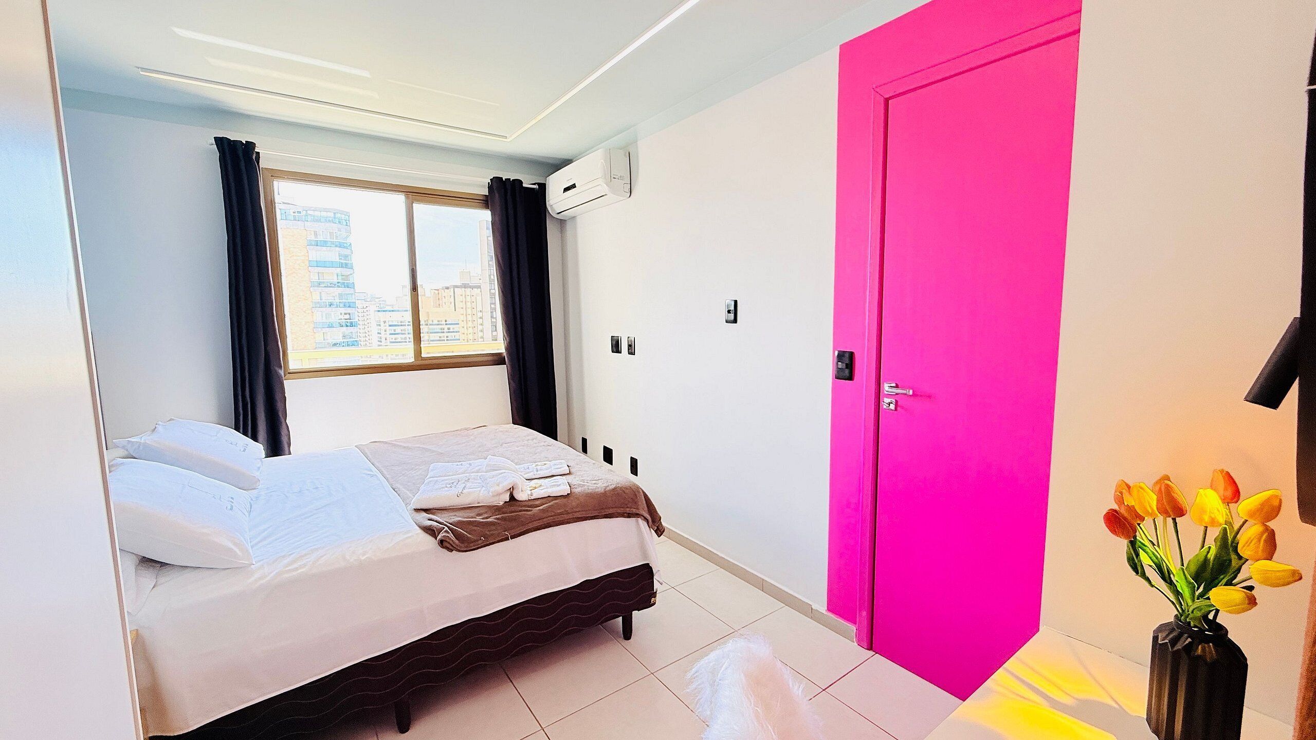 A hotel room with a bed, a window with curtains, an air conditioning unit, a bright pink door, and a vase with flowers.