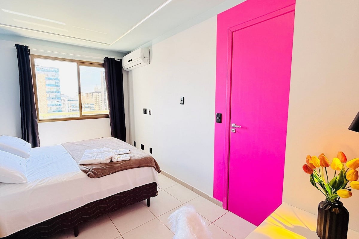 A hotel room with a bed, a window with curtains, an air conditioning unit, a bright pink door, and a vase with flowers.