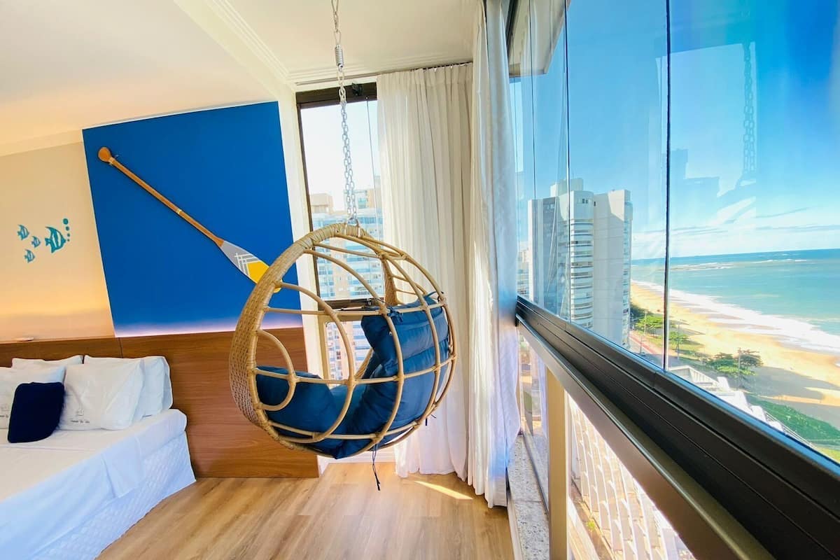 A bedroom with a hanging chair, a bed, a view of the beach, and a cityscape.