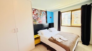 Habitación de hotel con una cama, un armario, una mesita de noche, una ventana con cortinas y una pared con un cartel colorido.