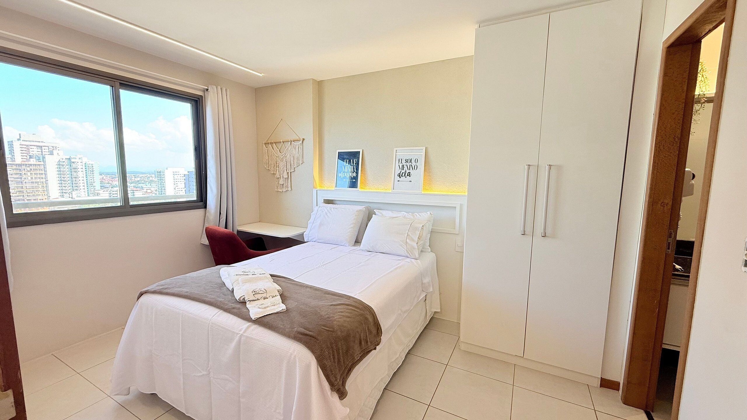 A hotel room with a bed, a desk, a wardrobe, and a view of the city.