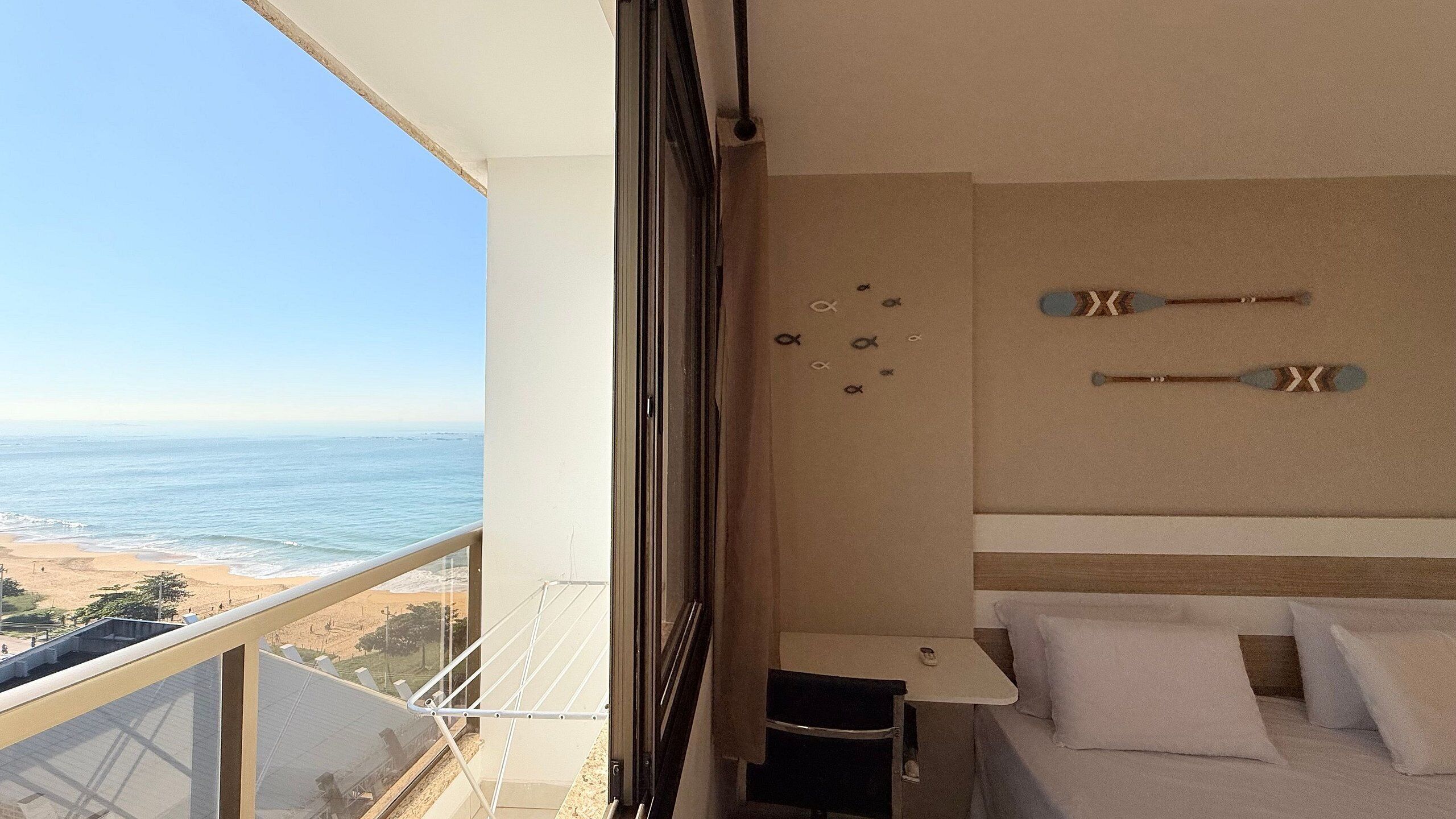 A balcony with a view of the beach and ocean, a bed with white bedding, and a wall with decorative items.