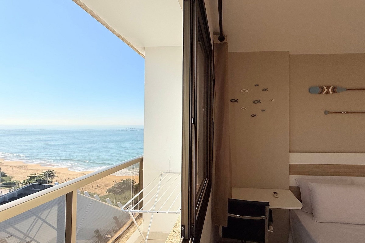 A balcony with a view of the beach and ocean, a bed with white bedding, and a wall with decorative items.