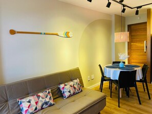 A modern living room with a sofa, patterned cushions, a dining table with chairs, and a wooden wall-mounted paddle.