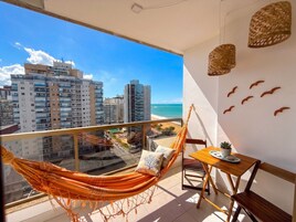 Un balcón con una hamaca, una mesa con un plato, y vistas a la ciudad y la playa.