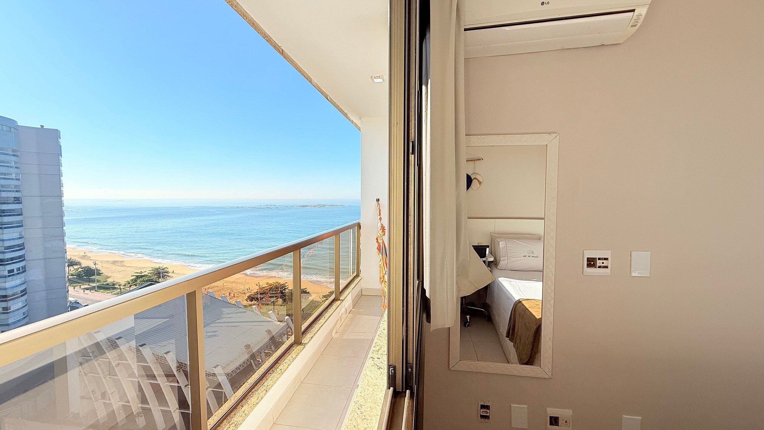 A balcony with a view of the beach and ocean, a bed with a white headboard, and a small window with a curtain.