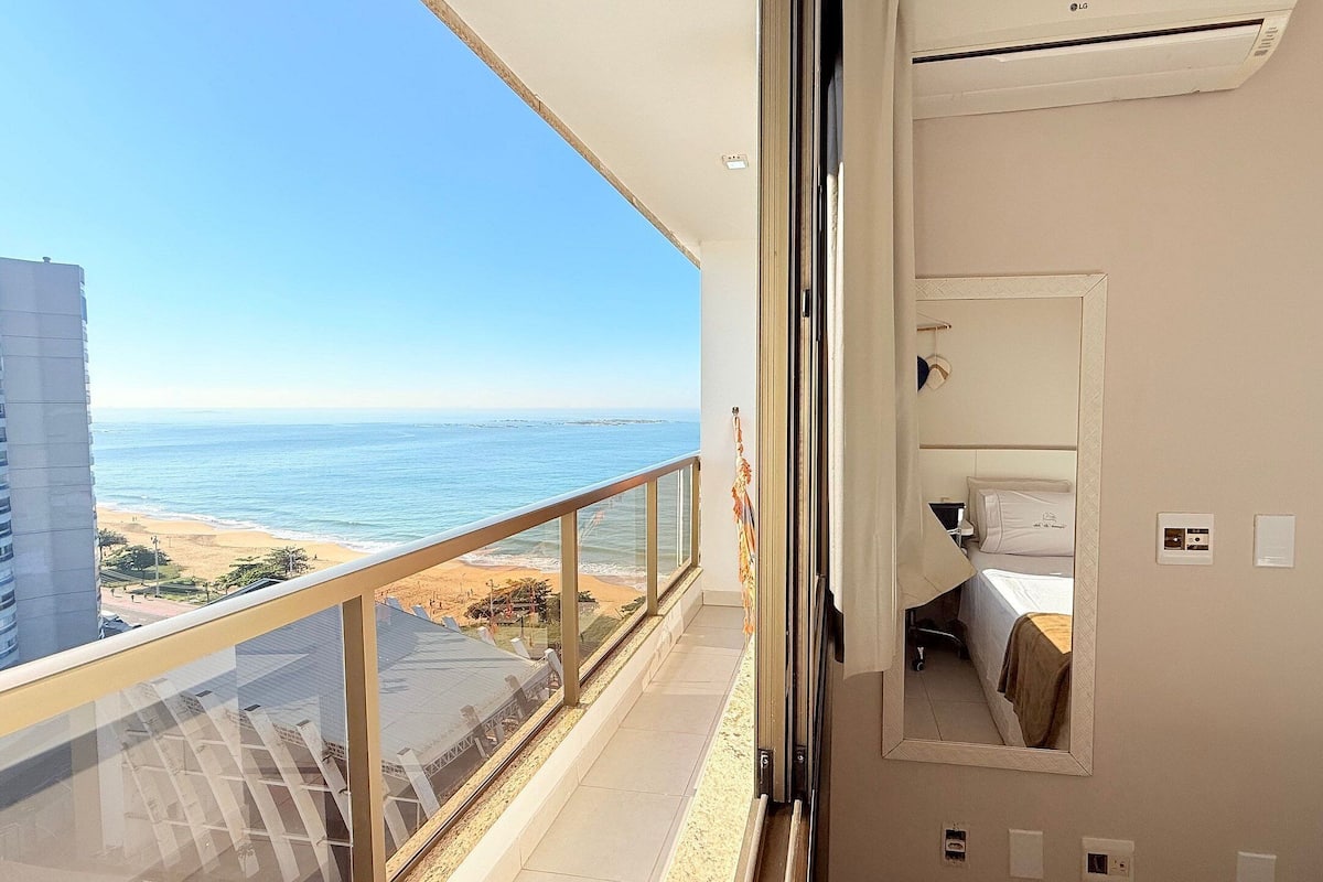 A balcony with a view of the beach and ocean, a bed with a white headboard, and a small window with a curtain.