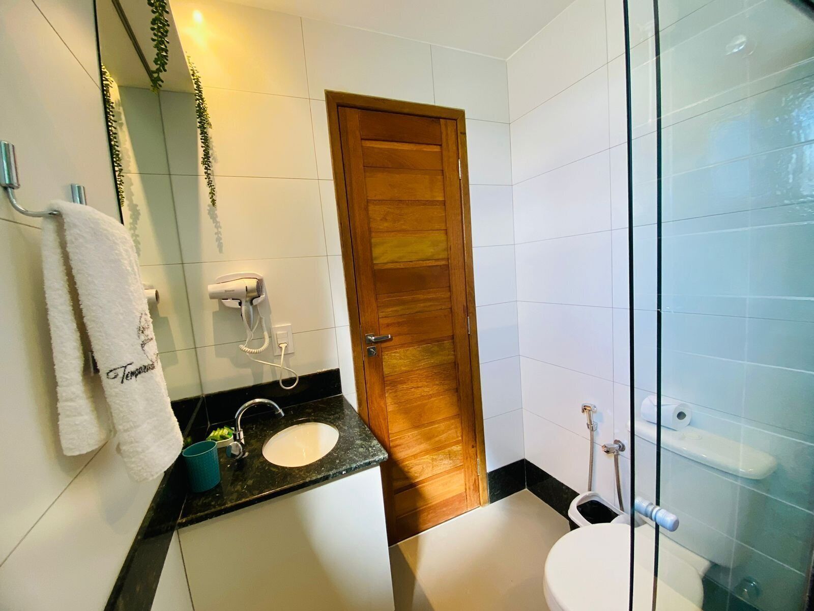 A bathroom with a wooden door, white tiles, a sink, a mirror, and a glass shower enclosure.