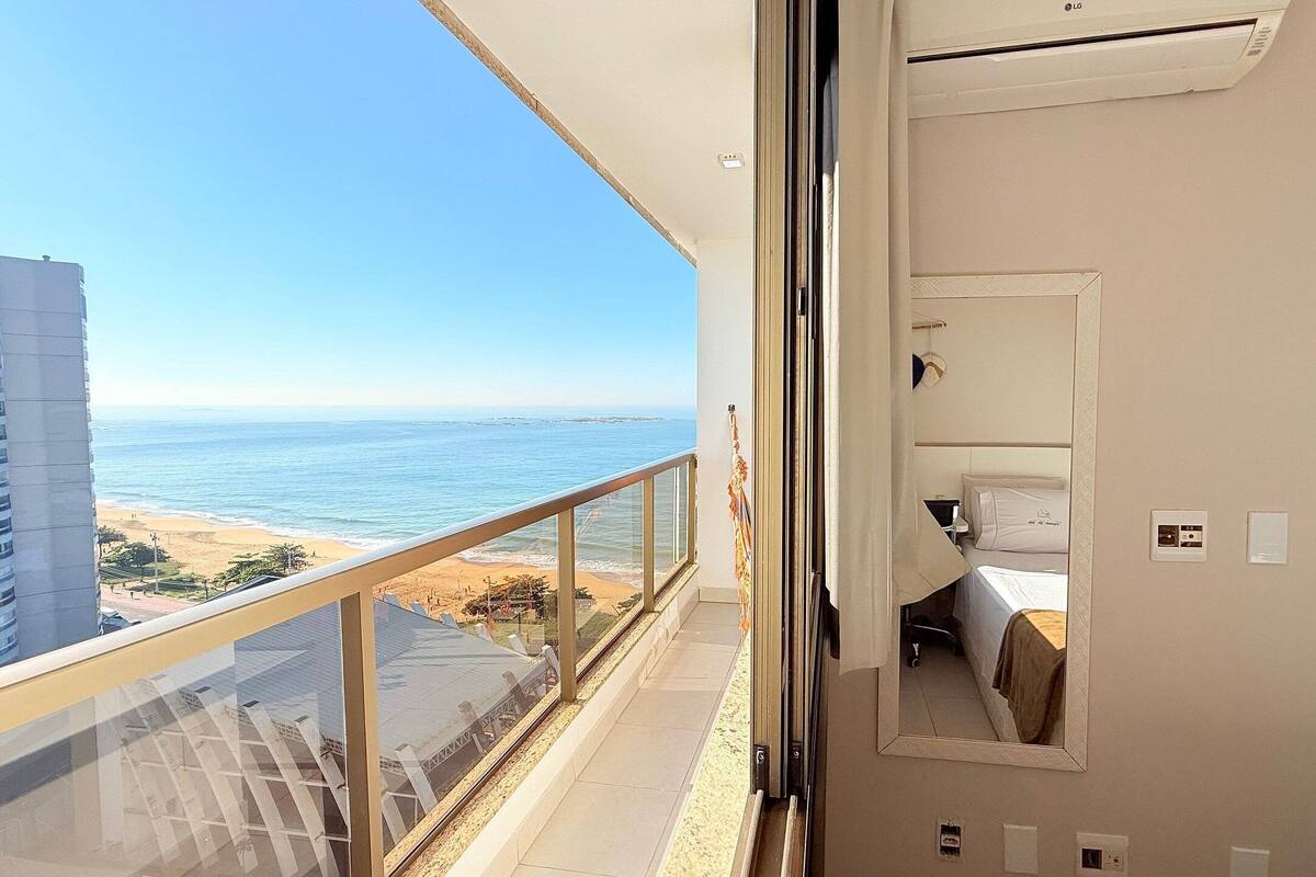 A balcony with a view of the beach and ocean, a bed with a white headboard, and a small window with a curtain.