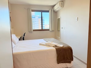 A hotel room with a bed, a window with curtains, an air conditioning unit, a mirror, and a bedside table. - Praia Dourada (Vila Velha)
