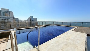 A rooftop infinity pool with a glass barrier, a clear view of the sea, and a cityscape in the background.